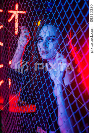 Caucasian woman in neon studio behind chain-link mesh.  89213380