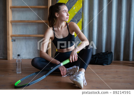 Portrait of female tennis player with racket sitting on gym floor Portrait of female tennis player with racket sitting on gym floor 89214591
