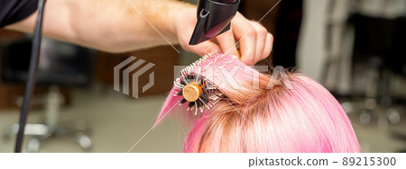 Drying short pink hair of young caucasian woman with a black hairdryer and black round brush by hands of a male hairdresser in a hair salon, close up Drying short pink hair of young caucasian woman with a black hairdryer and black round brush by hands of a male hairdresser in a hair salon, close up 89215300