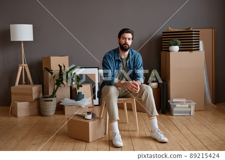 Horizontal shot portrait of young Caucasian man with beard on face sitting in loft room with stuff packed for moving to new apartment Horizontal shot portrait of young Caucasian man with beard on face sitting in loft room with stuff packed for moving to new apartment 89215424