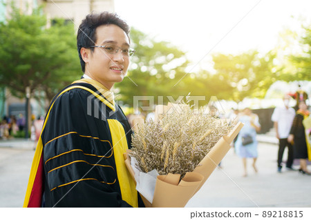 Happy Asian graduate student holding a beautiful bouquet of flowers in the university graduation ceremony. Master degree student in gown suit showing a bouquet of flower for photography. 89218815