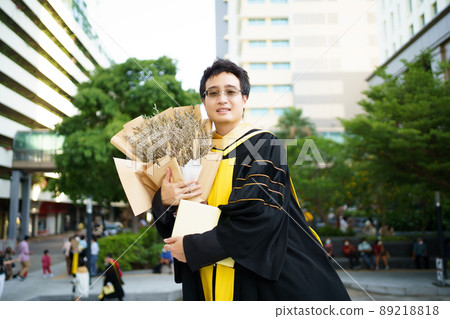 Happy Asian graduate student holding a beautiful bouquet of flowers in the university graduation ceremony. Master degree student in gown suit showing a bouquet of flower for photography. 89218818