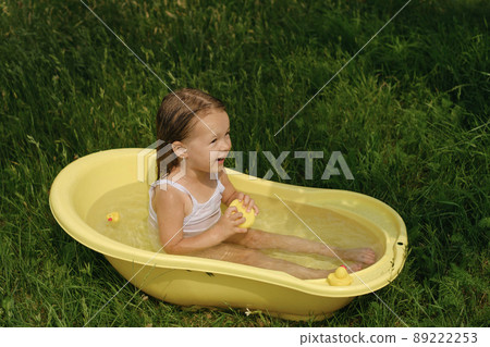 A girl bathes in water in a small tub in nature outdoors and plays with toy ducks. An infant baby takes a bath and laughs merrily 89222253