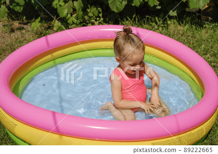 A cute little girl in a swimsuit splashes in the water in a small multicolored inflatable swimming pool. A hot sunny summer day. A cute little girl in a swimsuit splashes in the water in a small multicolored inflatable swimming pool. A hot sunny summer day. 89222665
