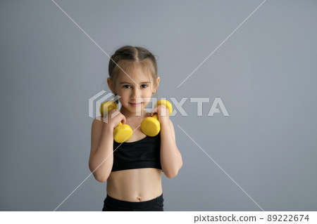 Cute beautiful sporty girl with braided pigtails plays fitness by lifting heavy dumbbells. Healthy, strong child in a sportswear . Person with sports equipment on a grey studio background copy space 89222674