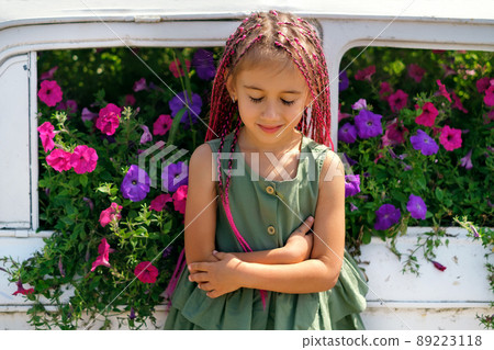 A girl with African pigtails in front of a flowerbed with petunias in a decorative car. The child with his eyes closed from inspiration and enjoyment of nature and the smell of flowers. zizi kanekalon 89223118