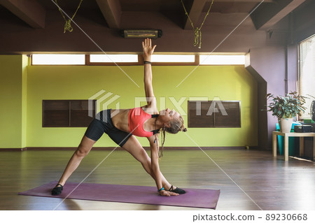 A woman practicing yoga, performs the exercise Utthita Trikonasana, the stretched triangle pose, stands on a mat in the studio 89230668