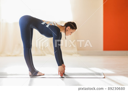 A beautiful woman does yoga, performs the Ardha uttanasana exercise, stands leaning forward on the mat, trains in blue sportswear in the studio A beautiful woman does yoga, performs the Ardha uttanasana exercise, stands leaning forward on the mat, trains in blue sportswear in the studio 89230680