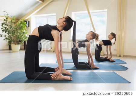 Young woman trainer with two little students perform ushtrasana exercise, camel pose. A group of athletes train in the studio on gymnastics mats 89230695