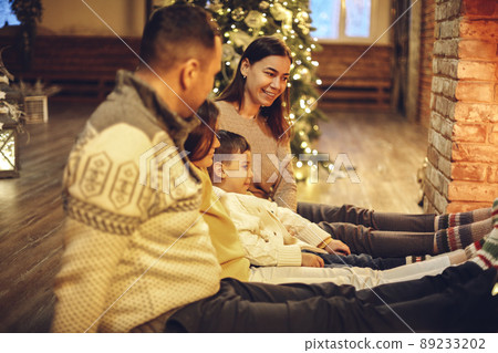 Happy young family parents with two small kids gathering around brick fireplace in cozy and warm country house during Christmas holidays Happy young family parents with two small kids gathering around brick fireplace in cozy and warm country house during Christmas holidays 89233202