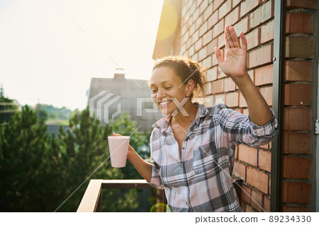 Pretty woman smiles and waves greeting, standing on balcony during coffee break 89234330