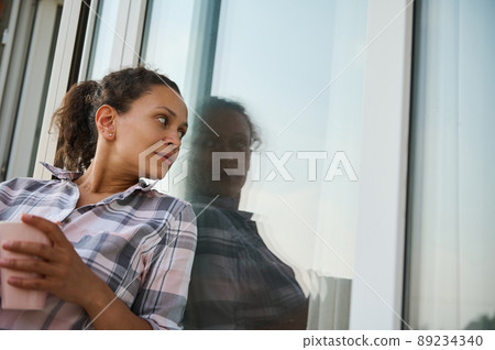 Charming woman with cup of coffee, looks at her reflection on the balcony window Charming woman with cup of coffee, looks at her reflection on the balcony window 89234340