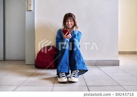 Teenage student in headphones with backpack smartphone sitting on floor near wall. 89243718