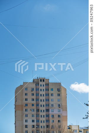 A tall apartment building against a blue sky. A group of wires crossed randomly in front of the house in the sky. White clouds. A tall apartment building against a blue sky. A group of wires crossed randomly in front of the house in the sky. White clouds. 89247963