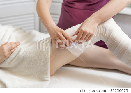 Beautician wraps body of woman in white bandage. Cosmetologist wraps the leg of the customer. Body shaping. Beautician wraps body of woman in white bandage. Cosmetologist wraps the leg of the customer. Body shaping. 89250075