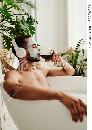 Young man in mask drinking red wine and listening to music in headphones while relaxing in bath 89250796