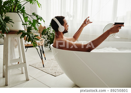 Young man relaxing in bathtub with glass of red wine and enjoying music in headphones during spa procedure in bathroom 89250801