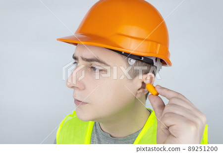 A worker in a hard hat and green vest puts on ear plugs. Industrial safety. Industrial noise protection. Compliance with the rules of conduct at loud work. Orange ear plugs 89251200