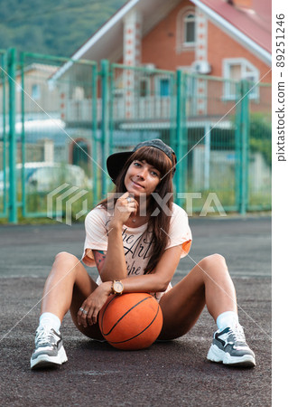 Beautiful young woman with tattoos, wearing a cap, sitting on the sport court with a basketball Beautiful young woman with tattoos, wearing a cap, sitting on the sport court with a basketball 89251246
