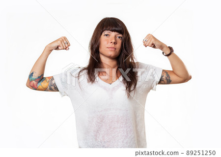 Concept of feminism and equal rights for women. Portrait of caucasian young woman with tattoo who shows her biceps. White background Concept of feminism and equal rights for women. Portrait of caucasian young woman with tattoo who shows her biceps. White background 89251250