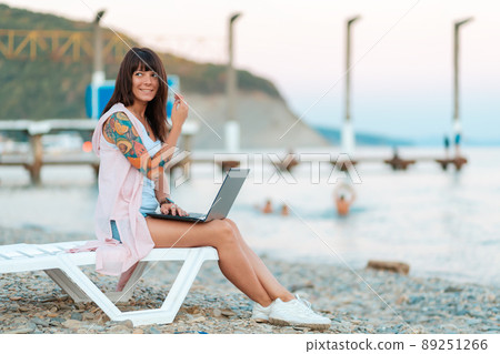 Summer vacation. A smiling woman with a tattoo on her arm sits on a chaise longue and works on a laptop. Side view. The concept of remote work and freelancing Summer vacation. A smiling woman with a tattoo on her arm sits on a chaise longue and works on a laptop. Side view. The concept of remote work and freelancing 89251266
