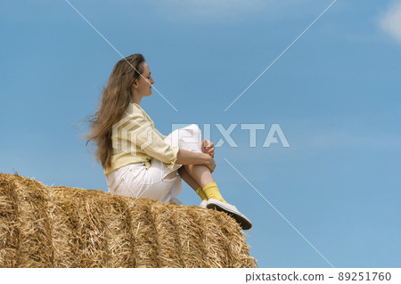 Girl with long blond hair sits thoughtfully on top of the haystack on blue sky background. Harvesting. Summer vacation. Girl with long blond hair sits thoughtfully on top of the haystack on blue sky background. Harvesting. Summer vacation. 89251760