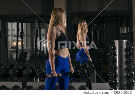 Young woman stands with dumbbells in hands in fitness club. Beautiful girl with sports figure is engaged in gym 89253068