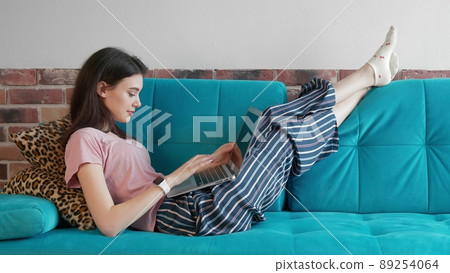 A young freelance woman in home clothes, working remotely in a home office, uses a laptop while lying on the couch. A modern woman browses the Internet in search of information, chatting online with A young freelance woman in home clothes, working remotely in a home office, uses a laptop while lying on the couch. A modern woman browses the Internet in search of information, chatting online with 89254064
