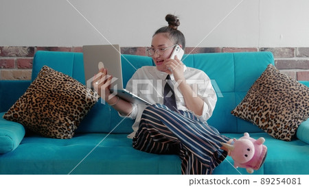A young freelance woman in a shirt and slippers, working remotely in a home office, uses a laptop while sitting on the couch. A modern woman is talking on the phone and typing on a laptop. 89254081