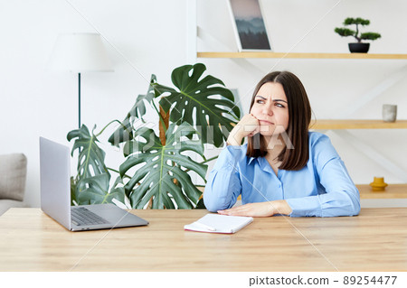 Young woman sitting at the table and working on laptop, upset woman. The girl rested her head and was tired from work, she was in a bad mood Young woman sitting at the table and working on laptop, upset woman. The girl rested her head and was tired from work, she was in a bad mood 89254477