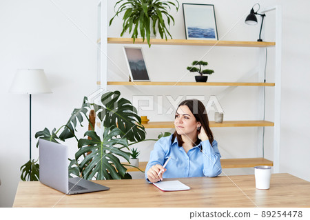 Video call. Young woman communicating via video call. She is sitting at the table and waving, while looks at laptop screen with smile 89254478