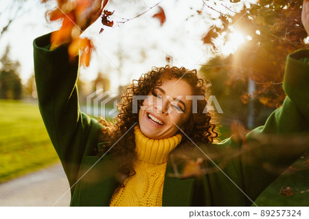 Smiling curly brunette covering her eyes with pleasure in autumn park touching hands with branches over her head 89257324