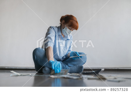 An adult woman in a medical mask on her face sits on the floor and counts money on a calculator, on a white background An adult woman in a medical mask on her face sits on the floor and counts money on a calculator, on a white background 89257328