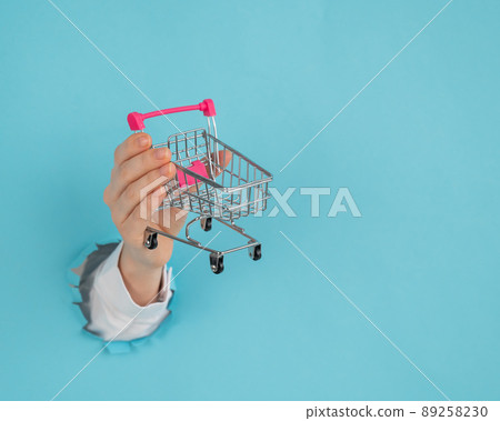 Woman's hand with a shopping cart on the palm sticking out of a hole in a paper blue background. Woman's hand with a shopping cart on the palm sticking out of a hole in a paper blue background. 89258230