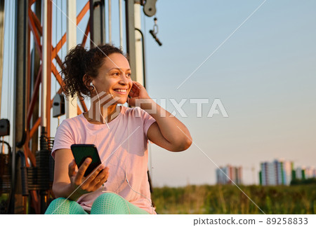 Cheerful multiethnic woman in sportswear, with earphones, listening to music, smiles looking aside while resting between sets during outdoor workout on sports field 89258833
