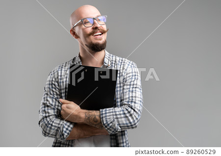 Smiling balded bearded man hold clipboard looking away from camera isolated on a grey background Smiling balded bearded man hold clipboard looking away from camera isolated on a grey background 89260529