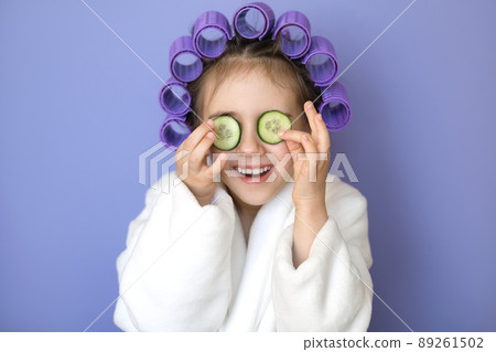 A humorous comical little beautiful girl with curlers in hair puts a slice of cucumber on eyes. Facial Skin Care, Moisturizing Natural Eye Mask. Loud children's laughter A humorous comical little beautiful girl with curlers in hair puts a slice of cucumber on eyes. Facial Skin Care, Moisturizing Natural Eye Mask. Loud children's laughter 89261502