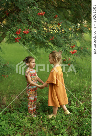 Children in the garden by a tree with rowan berries holding hands looking at each other. Fun outdoors during the summer holidays. Bright orange dresses of children on background of bright green grass Children in the garden by a tree with rowan berries holding hands looking at each other. Fun outdoors during the summer holidays. Bright orange dresses of children on background of bright green grass 89261503