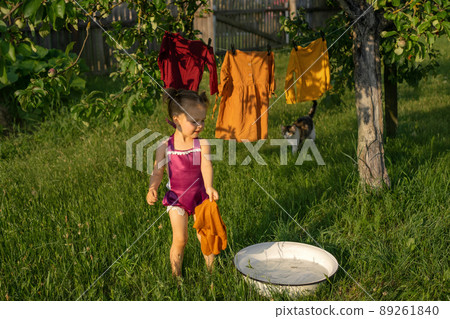 A cute girl splashes in a basin with soapy water, washes clothes and hangs clothes for drying in the garden. Washing and drying of colored clothes in the outdoors. The Little Helper and his cat 89261840