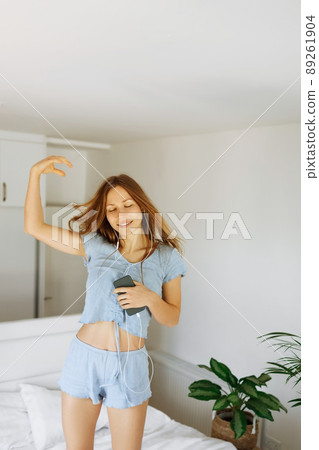 Young teen girl in pajama standing on her bed and singing a song. Cheerful caucasian adolescent girl with headphones dancing on her bedroom .  89261904