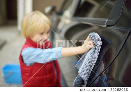 Little boy washing black car near garage. Child having fun and helping his parents. Family upbringing. Focus on the napkin 89262018