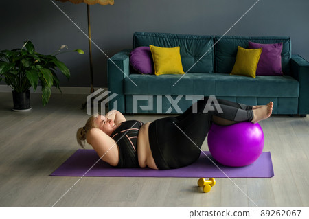 Fat chubby woman does Exercise for lifting the trunk at home in the hallway on the floor, putting her feet on a fit ball. An obese lady does sports lying on a fitness mat. A strong woman. Fat chubby woman does Exercise for lifting the trunk at home in the hallway on the floor, putting her feet on a fit ball. An obese lady does sports lying on a fitness mat. A strong woman. 89262067