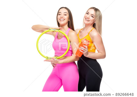Two beautiful, athletic, slim and cheerful female friends are smiling and holding a hoop and a bottle of water in their hands. Lifestyle concept with sports and gym. Isolated on white background. 89266648