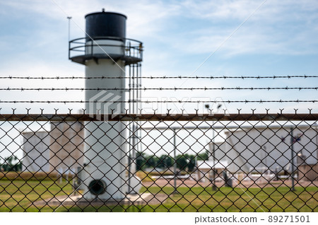 chemical facility security chain fence with barbed wire top in focus and a blurred tank farm and emergency flare in the background 89271501