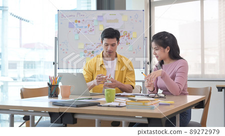 Happy young female employee discussing online project with her colleague at creative office. Happy young female employee discussing online project with her colleague at creative office. 89272979