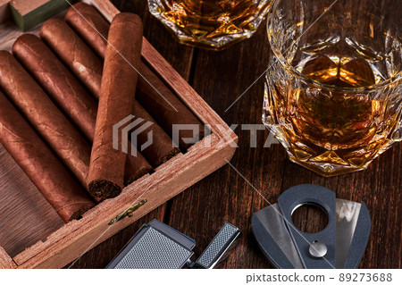 Box with cuban cigars, lighter and cutter on old wooden table top. Two glasses of whiskey or alcohol on the background. 89273688