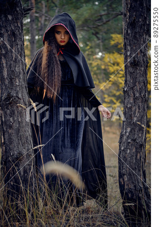 Long-haired witch in black, long dress with cape and hood. Posing in pine forest among trees. Spells, magic and witchcraft. Full length portrait. 89275550
