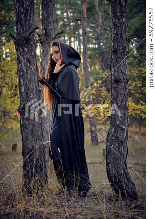 Long-haired witch in black, long dress with cape and hood. Posing in pine forest among trees. Spells, magic and witchcraft. Full length portrait. 89275552