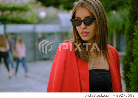 Blonde girl in a red lady-type pantsuit and black top, pendant and sunglasses is walking alone in the city. Fashion and style. Close-up. Blonde girl in a red lady-type pantsuit and black top, pendant and sunglasses is walking alone in the city. Fashion and style. Close-up. 89275592