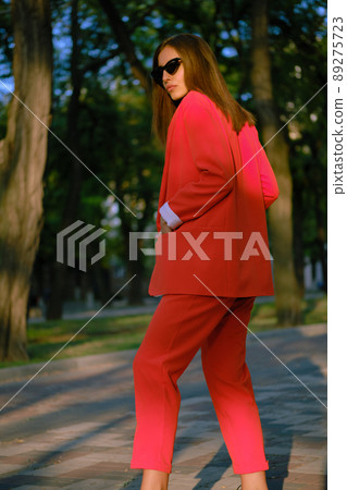 Blonde girl in a red lady-type pantsuit and black top, watch, ring, pendant and sunglasses is walking alone in the city. Fashion and style. Close-up. Blonde girl in a red lady-type pantsuit and black top, watch, ring, pendant and sunglasses is walking alone in the city. Fashion and style. Close-up. 89275723